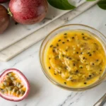 Fresh passion fruit puree in a glass bowl, surrounded by halved passion fruits on a white marble countertop.